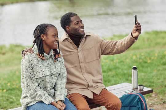 Happy African couple smiling at camera making selfie portrait on mobile phone while sitting on bench in the park