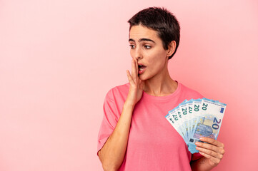 Young caucasian woman holding banknotes isolated on pink background is saying a secret hot braking news and looking aside