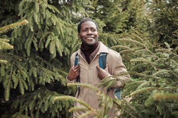 African happy man with backpack walking along fir trees in the park outdoors