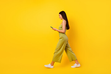 Fototapeta na wymiar Full length profile photo of gorgeous positive filipino girl hold telephone texting walking isolated on yellow color background