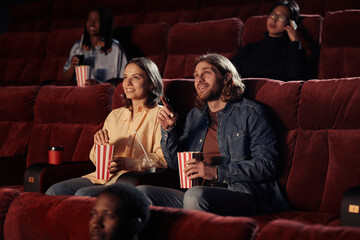 Young couple eating popcorn and watching movie together on armchairs in the cinema - obrazy, fototapety, plakaty