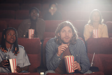 Young guy with popcorn concentrating on watching movie together with his friends in the cinema