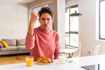 Young mixed race man having breakfast in a kitchen on the morning showing fist to camera, aggressive facial expression.