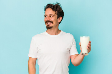 Young caucasian man holding a glass of milk isolated on blue background looks aside smiling, cheerful and pleasant.