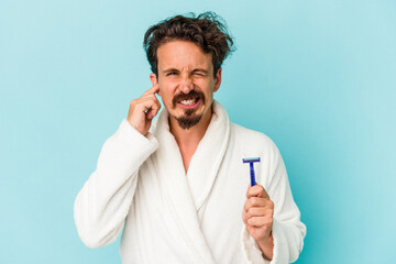 Young caucasian man holding a razor blade isolated on blue background covering ears with hands.