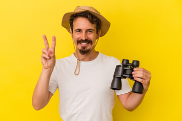 Young caucasian man holding binoculars isolated on yellow background showing number two with fingers.