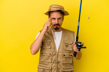 Young caucasian fisherman holding rod isolated on yellow background pointing temple with finger, thinking, focused on a task.
