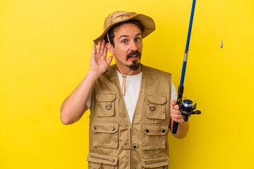 Young caucasian fisherman holding rod isolated on yellow background trying to listening a gossip.