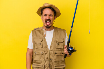 Young caucasian fisherman holding rod isolated on yellow background screaming very angry and aggressive.