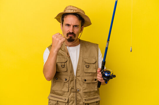 Young caucasian fisherman holding rod isolated on yellow background showing fist to camera, aggressive facial expression.