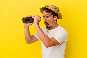 Young caucasian man holding binoculars isolated on yellow background