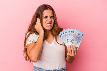 Young caucasian woman holding bills isolated on pink background  covering ears with hands.