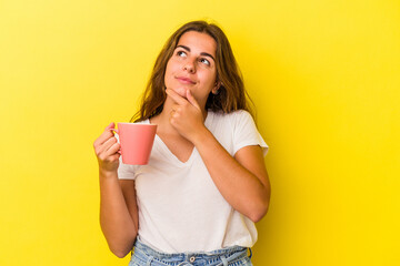 Young caucasian woman holding a mug isolated on yellow background  looking sideways with doubtful and skeptical expression.