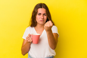 Young caucasian woman holding a mug isolated on yellow background  showing fist to camera, aggressive facial expression.