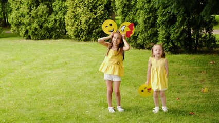 Two funny little girls are making faces and dancing in the park on the lawn, holding different emoticons in hands