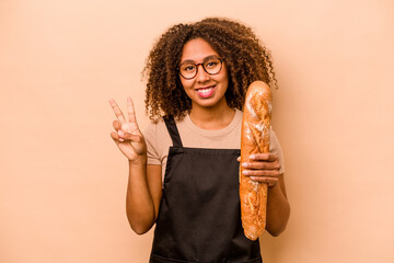 Young baker African woman holding a loaf of bread isolated on beige background showing number two...