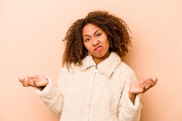 Young African American woman isolated on beige background doubting and shrugging shoulders in...