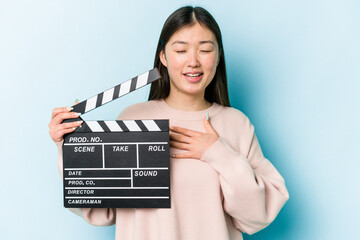 Young asian woman holding clapperboard isolated on blue background laughs out loudly keeping hand...