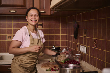 Multiethnic cute woman housewife stands at the kitchen countertop with sterilize glass jars for filling with cherry jam