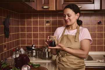 Cheerful African American housewife holds a cherry jam jar with the burlap on the lid, standing at home kitchen interior