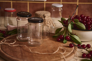 Close-up of sterile jars with lids on wooden board next to a bowl with freshly picked cherries on...