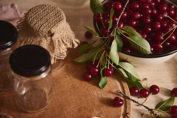 Top view of sterile jars with lids on wooden board next to a bowl with freshly picked cherries on the kitchen countertop