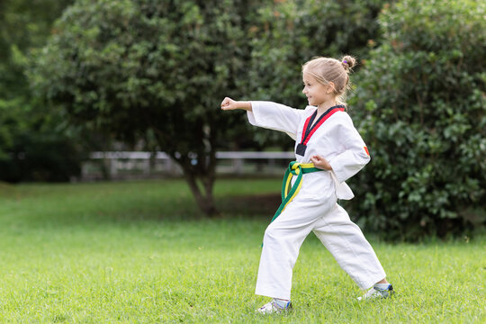 Caucasian little girl seven years old in kimono with yellow green belt exercising Taekwondo at summer park alone during coronavirus covid-19 lockdown, self isolation and social distancing