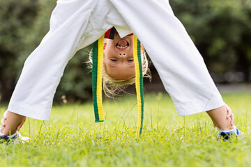 Caucasian little girl seven years old in kimono with yellow green belt exercising Taekwondo at summer park alone during coronavirus covid-19 lockdown, self isolation and social distancing © Алина Бузунова