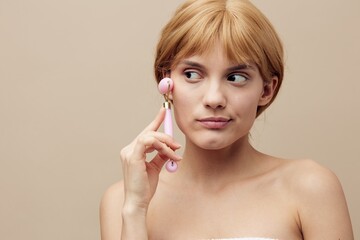 Close-up photo. An emotional woman in a towel with clean, beautiful, well-groomed skin, short red hair on a beige background does a facial massage with a new massage roller. Horizontal studio shot.