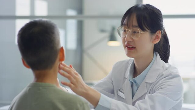 Asian female doctor examining throat and neck of tween boy during health checkup in medical office
