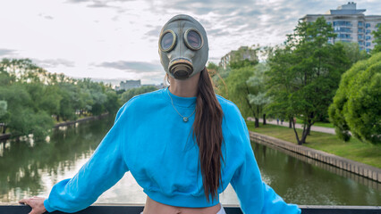 A girl in a gas mask stands on a bridge across the river. Alienation in the city. Fashion, ecology and loneliness concepts.
