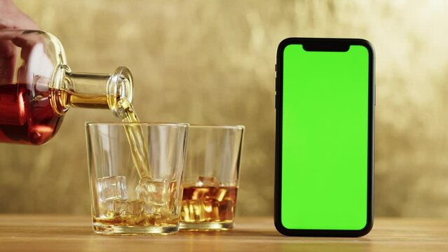 Brandy or whiskey close-up. Luxury cognac and smartphone with green chroma key screen. Alcohol amber drink with ice, drinking rum, liqour beverage in glass.