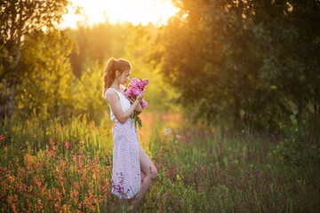 Beautiful happy young girl with peonies in nature outdoors in the sun. Beautiful woman in a field with flowers in a long skirt