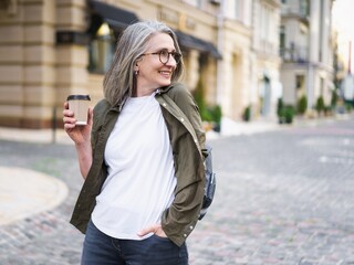 Businesswoman hold disposable cup with hot coffee. Mature woman spend free time traveling in european old town enjoying cup of tea, coffee. Grey haired woman in casual with backpack