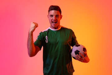 Portrait of happy caucasian male soccer player raising hand with football over neon pink lighting