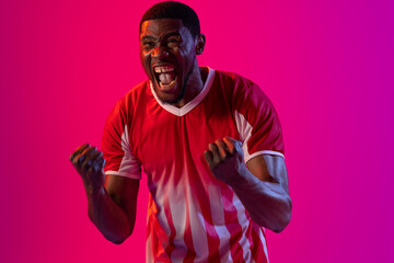 Happy african american male soccer player raising hands over neon pink lighting