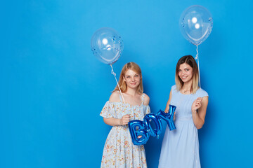 Two smiling girls in light summer dress, holding blue balloons and balloon with the inscription boy, isolated blue background.