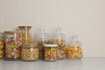 Glass containers with different breakfast cereals on white countertop near brick wall. Space for text