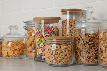 Glass containers with different breakfast cereals on white countertop near brick wall