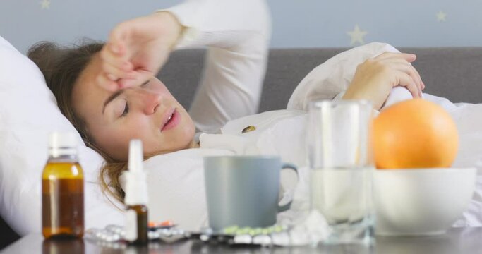 A young woman with bad cold lying in bed under the blanket and using nasal drops after blowing snot into a napkin. Woman using nose spray