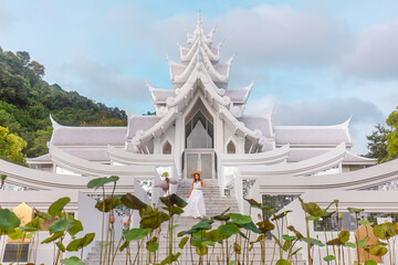 Traveler woman in dress walking in front of white and silver temple in thai style. Travel and vacation in Asia. Buddhist temple on Phuket Island in Thailand with blooming lotus in foreground. - obrazy, fototapety, plakaty
