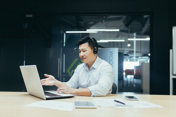 Angry and annoyed asian businessman with headset for video call, talking in online meeting, looking...