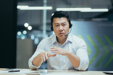 Serious and disappointed asian businessman with headset for video call, looking at web camera,...