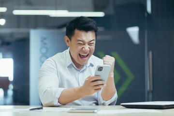 Happy and successful Asian businessman working in office, typing on phone and reading good news, celebrating triumph and victory