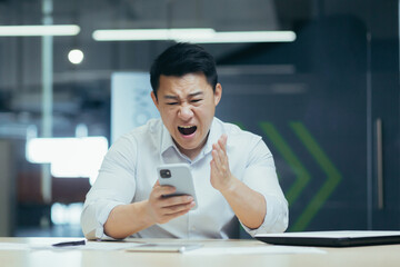 Angry businessman in office yelling on the phone, Asian man frustrated with received email.