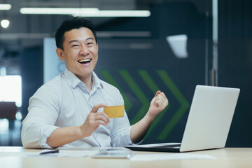 Happy Asian businessman smiling and looking at camera, holding bank credit card for purchase in...