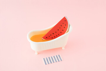 Watermelon slice in white bathtub and striped bath towel on the floor. Pink background.