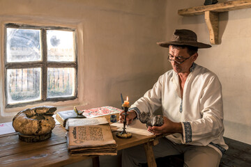 a man in old peasant clothes inside his dilapidated old house is sitting writing a letter at a table by candlelight and reading books