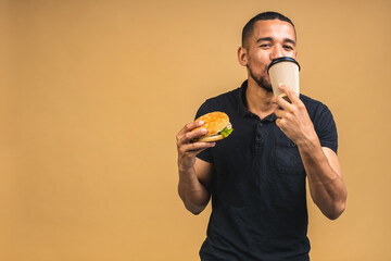 Hungry young african american black man eating hamburger isolated over beige background. Diet or Fast food concept. Drinking coffee. - 516419924
