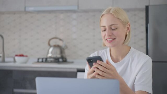 Cheerful blonde woman says bye on phone call when sitting at laptop in kitchen